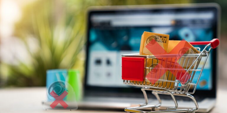 Un pequeño carrito de compras lleno de paquetes con pegatinas rojas de prohibido, frente a un portátil desenfocado que muestra una página web de compras. En primer plano a la izquierda, un lector de tarjetas con una X roja también está presente.